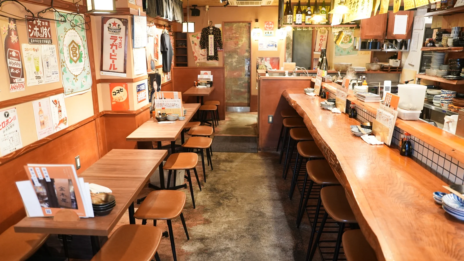 昭和のポスターが並ぶ壁と木のテーブル、長いカウンターを見渡す店内